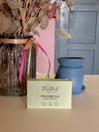A box of The Natural Beauty Pot "The First Cut wax melts' sits on a tabletop in a styled interior scene with dried flowers in a glass jar, a blue ceramic wax melt burner, and a pink wall in the background.
