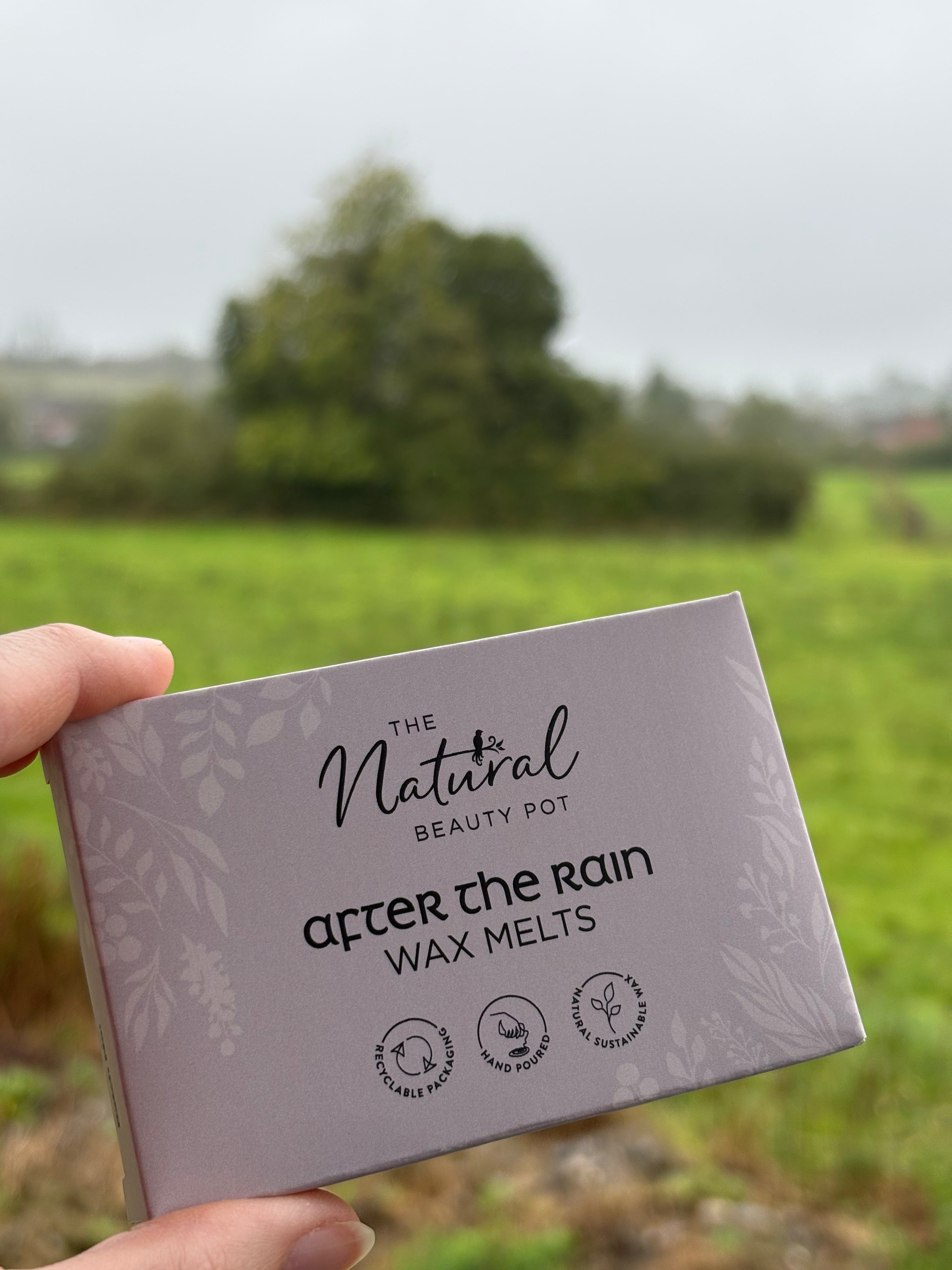 A hand holds a square box of 'The Natural Beauty Pot' after the rain wax melts, with a blurred green field and trees in the background.