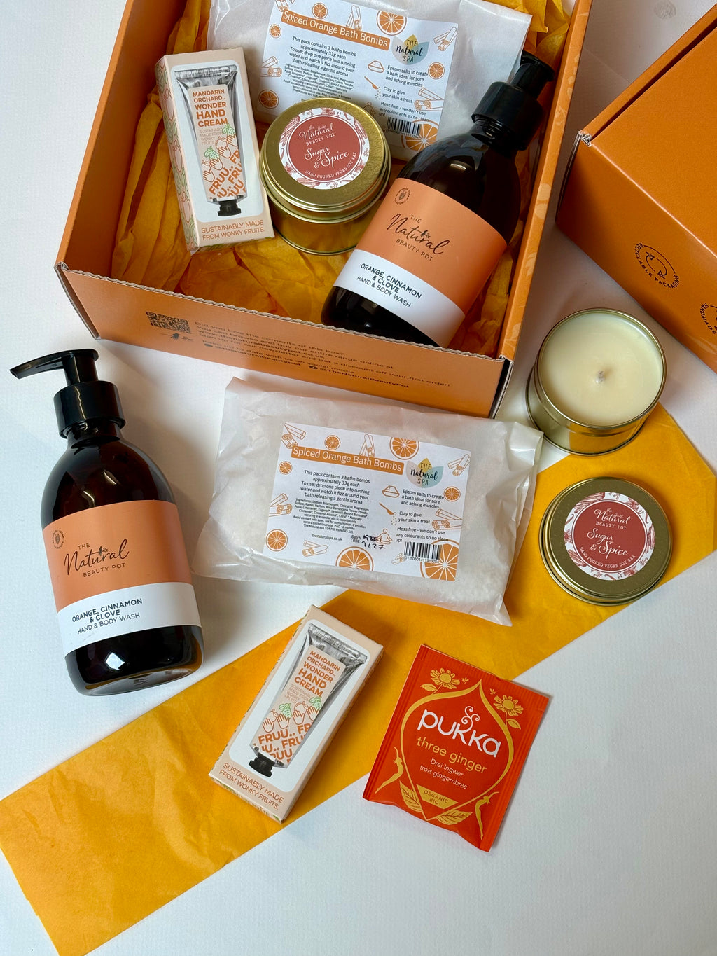 An open orange gift box filled with bath and body items, including amber bottle of handwash in orange, cinnamon and clove, a candle, spiced orange bath bombs and herbal tea arranged on a white surface with additional matching tissue around it.
