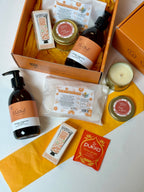 An open orange gift box filled with bath and body items, including amber bottle of handwash in orange, cinnamon and clove, a candle, spiced orange bath bombs and herbal tea arranged on a white surface with additional matching tissue around it.