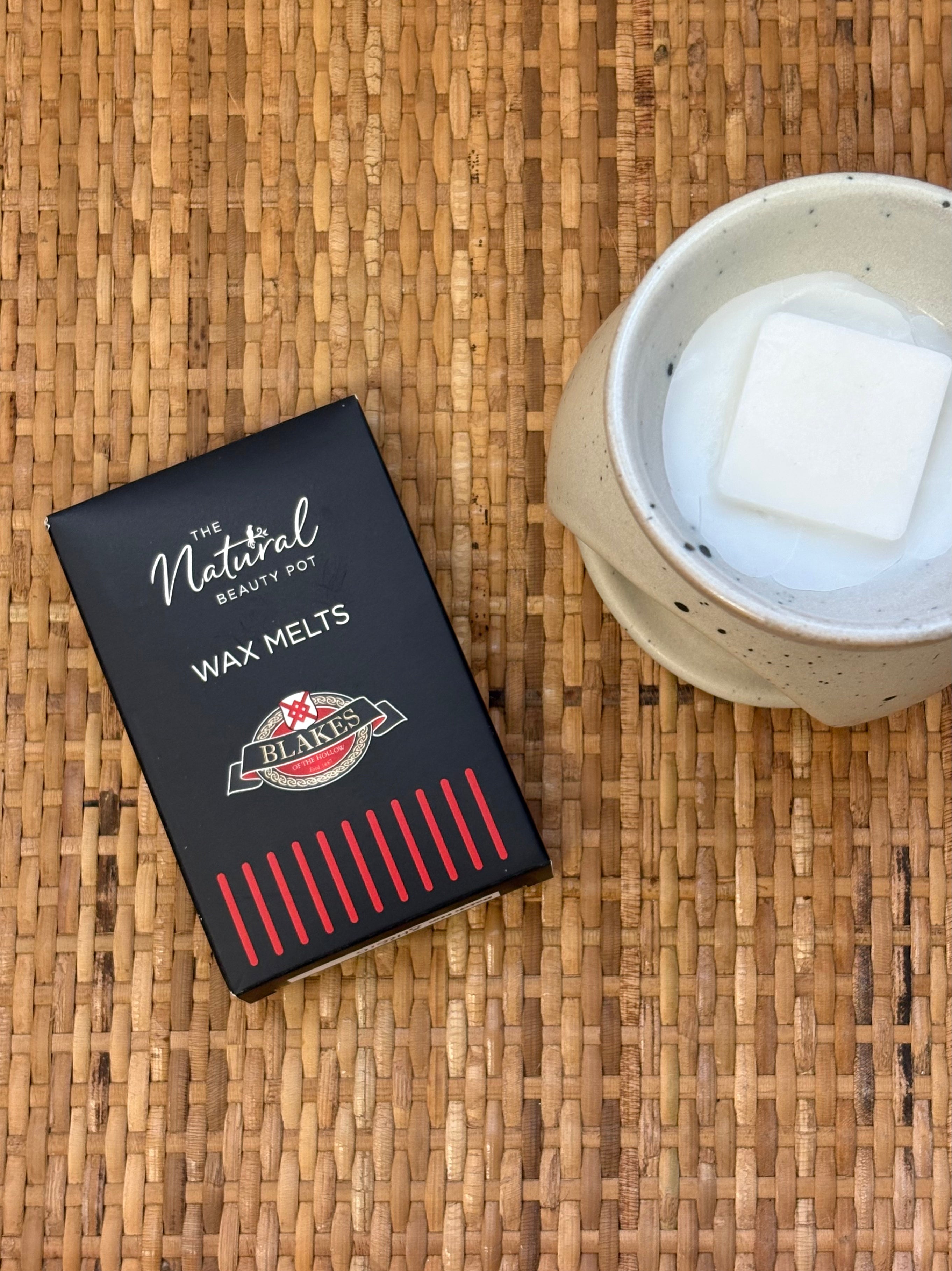 A black box of The Natural Beauty Pot Blakes of the Hollow Wax Melts placed on a woven tan mat next to a speckled ceramic mug containing a white wax melt cube.