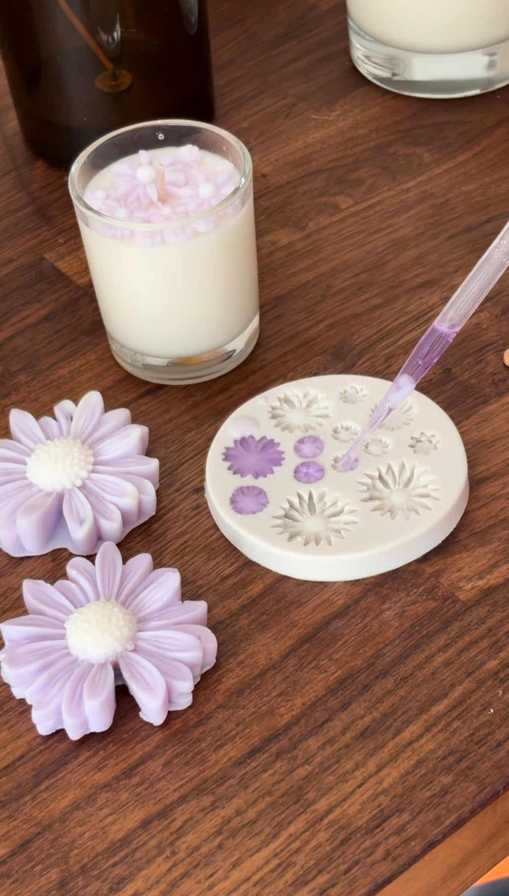 a workshop to make floral patterns in wax in lilac, with a pipette applying more wax. Two finished lavender flower-shaped wax melts and a finished candle with wax flowers on top