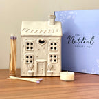 Ceramic two story house-shaped tea light holder with candles on a wooden surface, featuring 'The Natural Beauty Pot' branding.