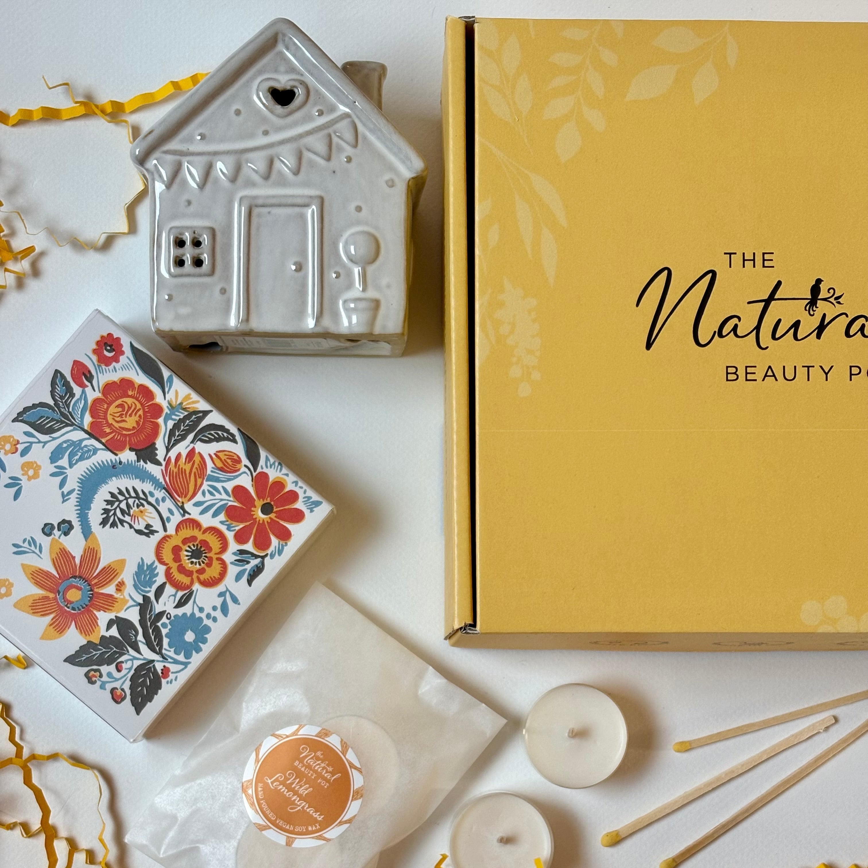 Ceramic house-shaped container, floral box, yellow box labeled 'The Natural Beauty Box', and candles on a white surface.