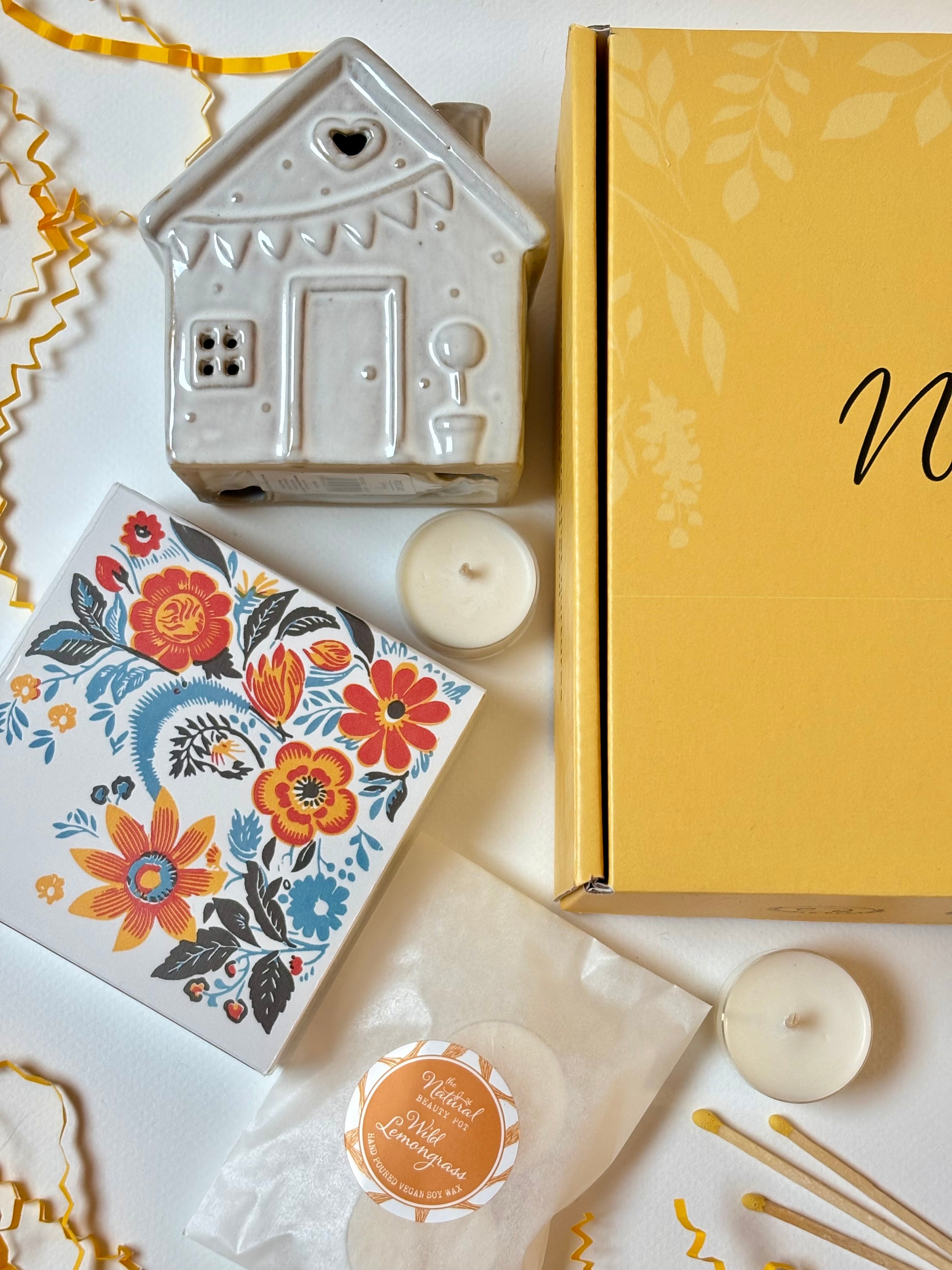 Cottage tea light house, floral matches box, and tea lights on a white surface with a yellow gift box in the background.