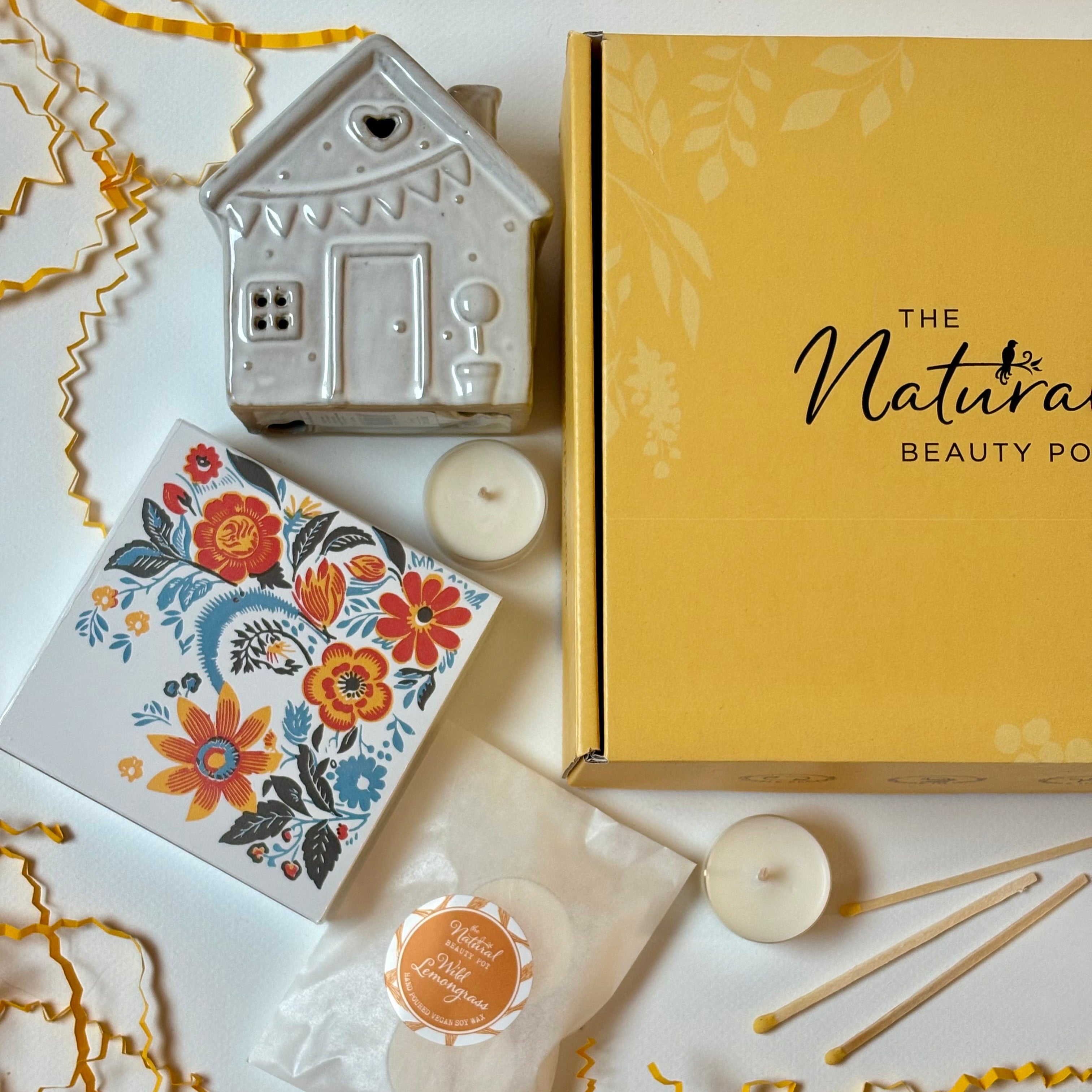 Decorative items including a ceramic house, floral matches box, and candles on a white surface with 'The Natura Beauty Pot' branding.