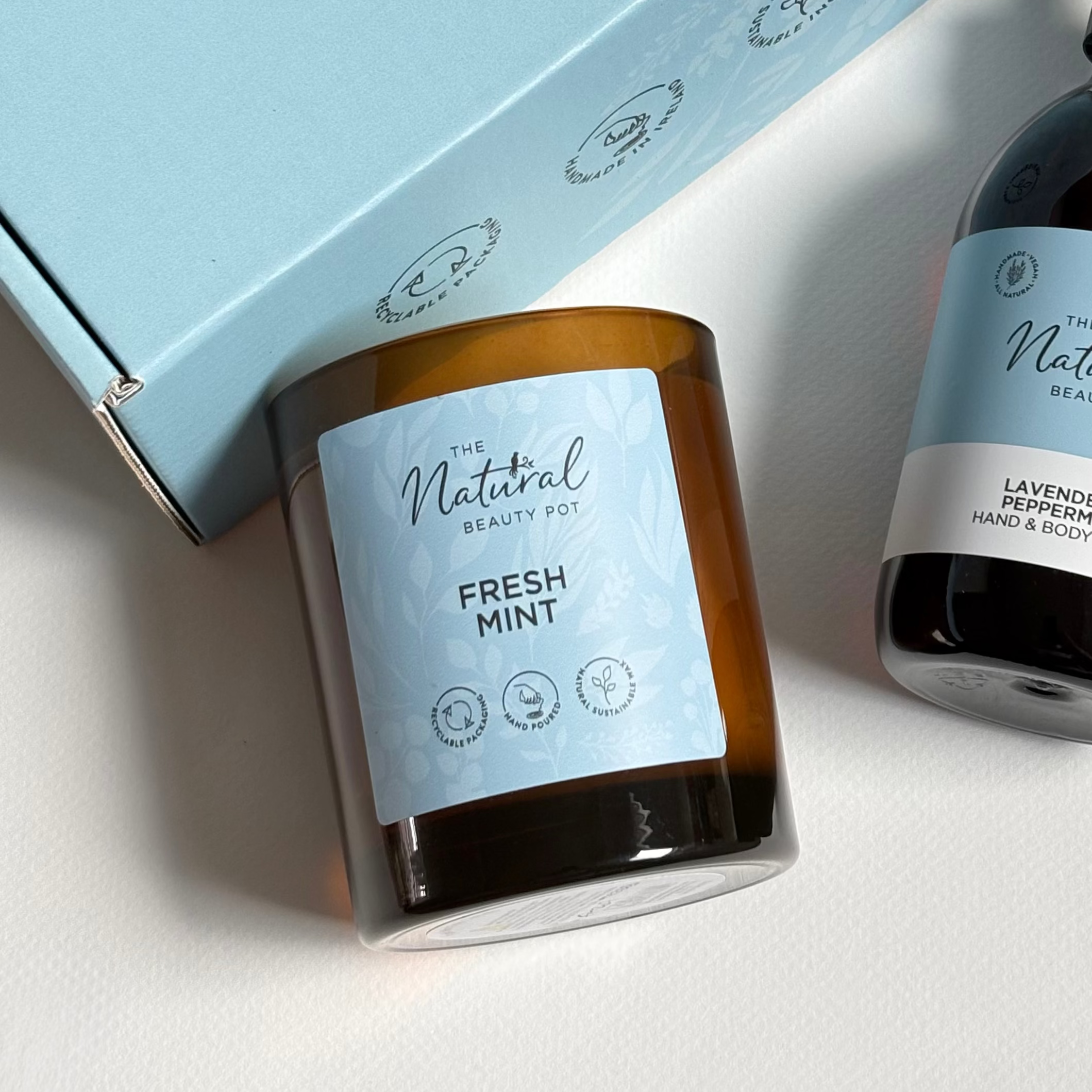 The Natural Beauty Pot gift set featuring a light blue box, a fresh mint scented candle in an amber glass jar, and a lavender and peppermint hand wash in an amber bottle with a black pump, arranged on a white surface.