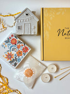 Decorative gift items including a ceramic house, matches in a floral box, and candles on a white surface.