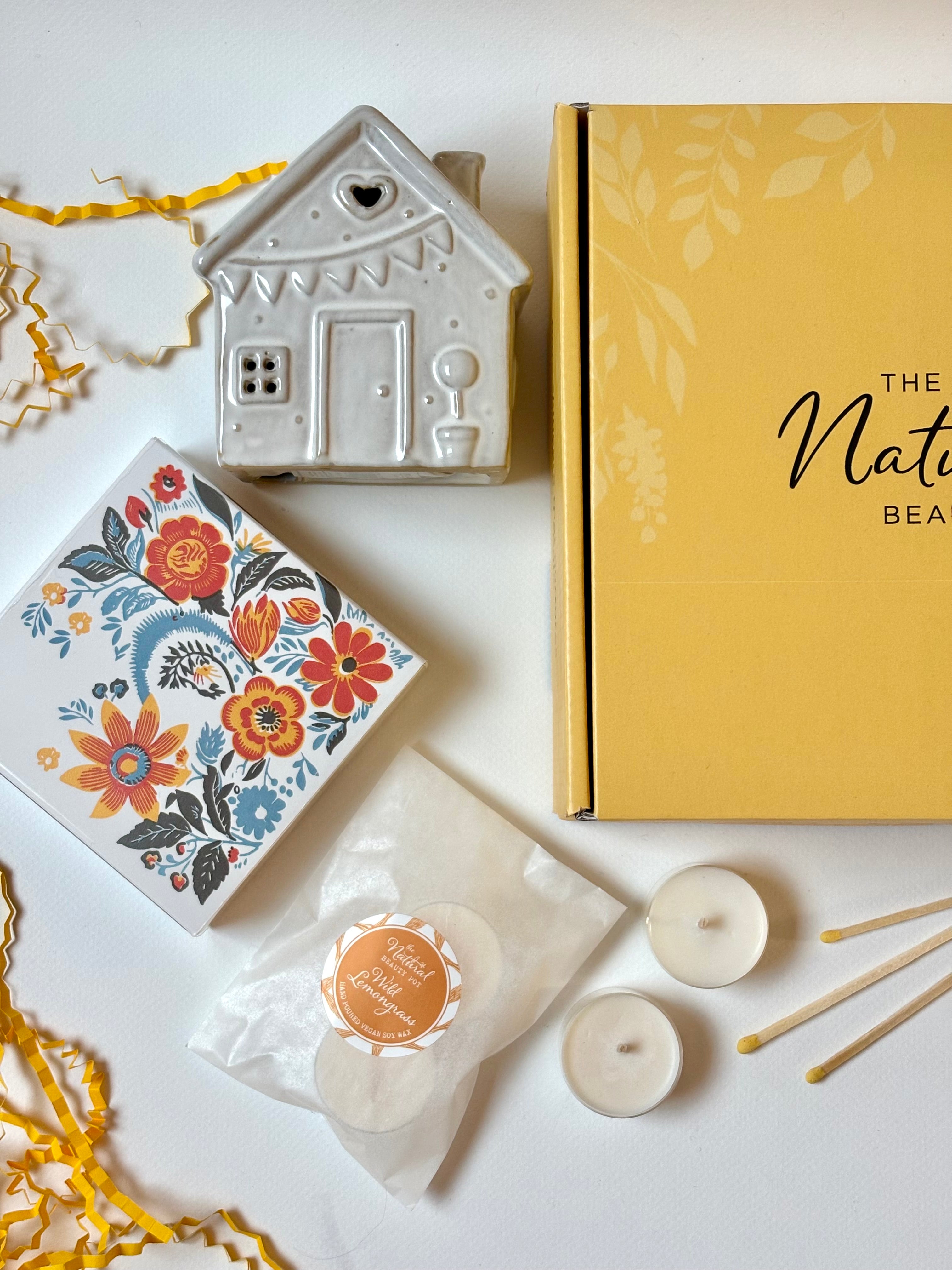 Decorative gift items including a ceramic house, matches in a floral box, and candles on a white surface.