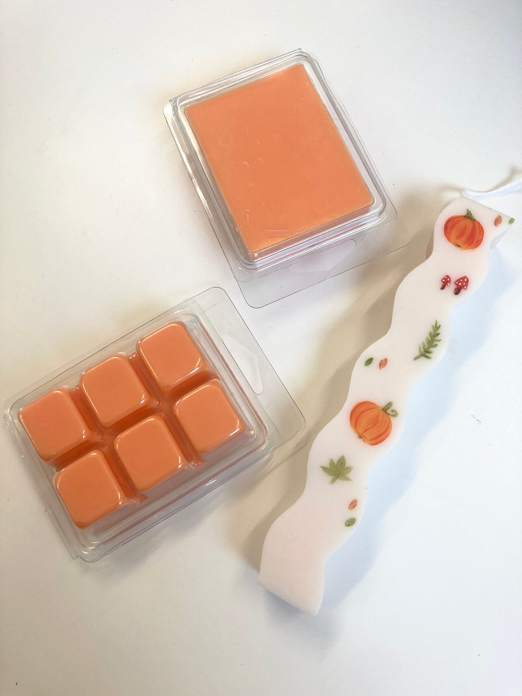 An orange wax melt set with a single square wax block in a clear clamshell and six smaller wax cubes alongside a white decorative candle with orange pumpkins and fall leaves.