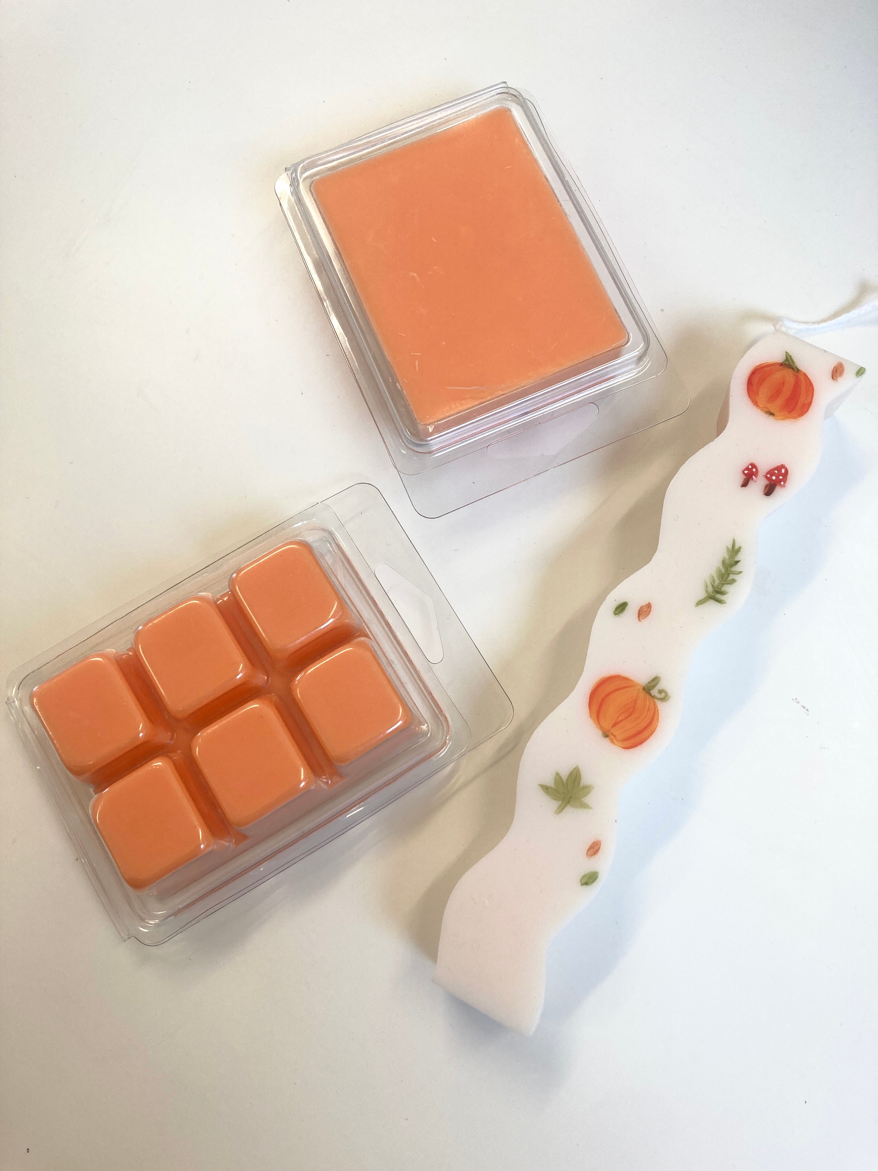 An orange wax melt set with a single square wax block in a clear clamshell and six smaller wax cubes alongside a white decorative candle with orange pumpkins and fall leaves.