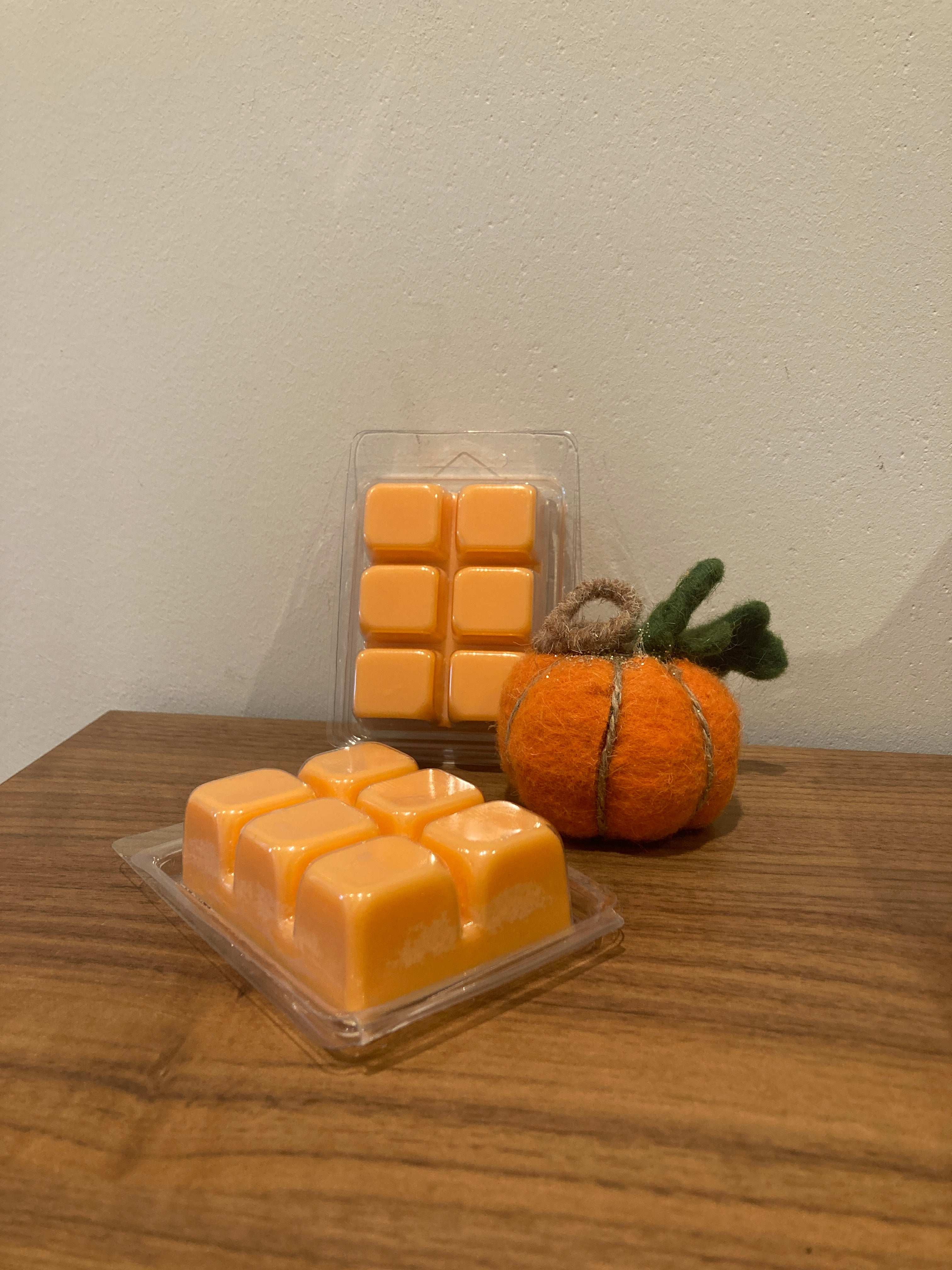 Two wax melts in clear plastic clamshell packaging, one shaped like a cube and the other like a rounded square, both in a bright orange color. They are styled on a wooden table next to a small felted orange pumpkin decoration against a plain beige wall.
