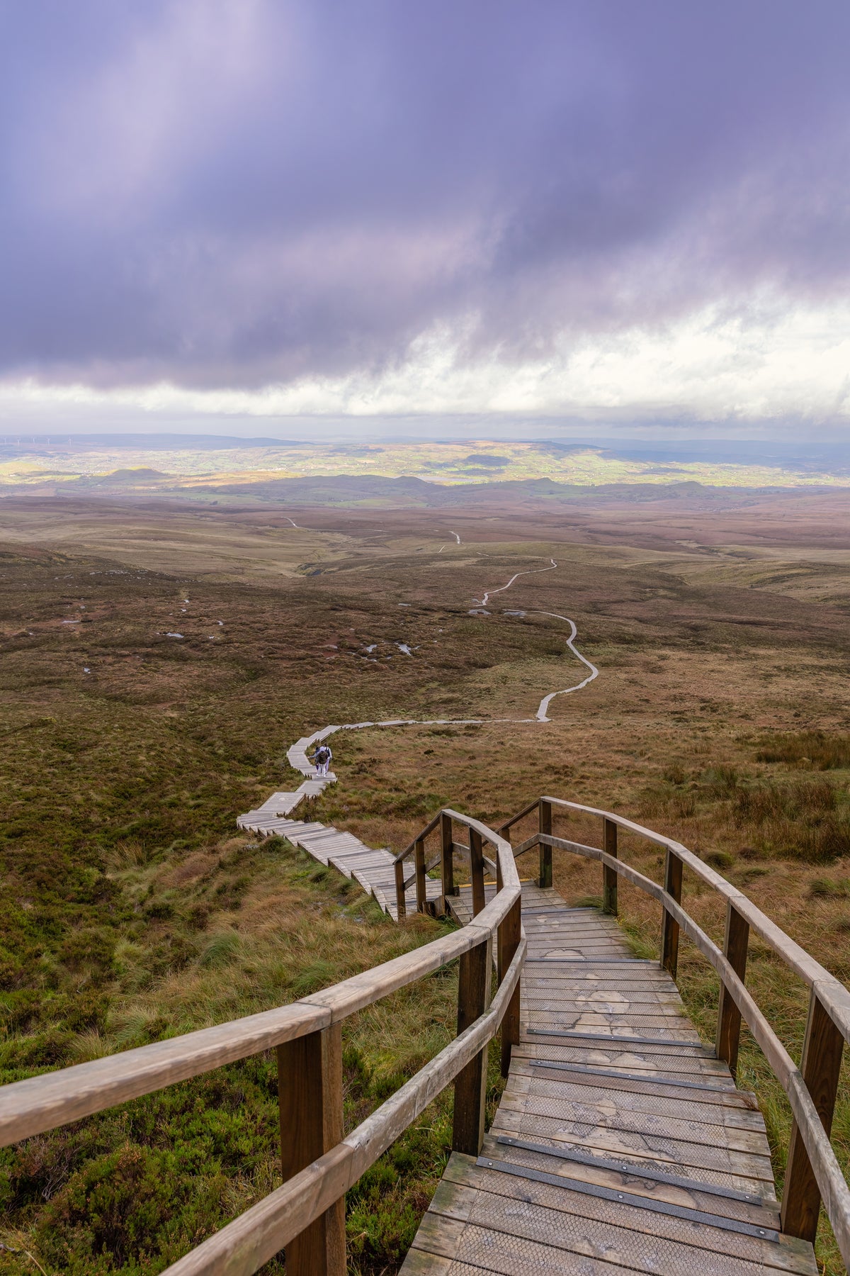A wooden boardwalk staircase curves through a vast moorland landscape under a dramatic cloudy sky, with rolling hills and winding paths below.