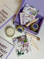 A lavender-themed gift set arranged on a light surface, featuring a purple box with candles, matchsticks, a hand cream tube. Visible text includes 'Happy Birthday' and 'Lavender'.