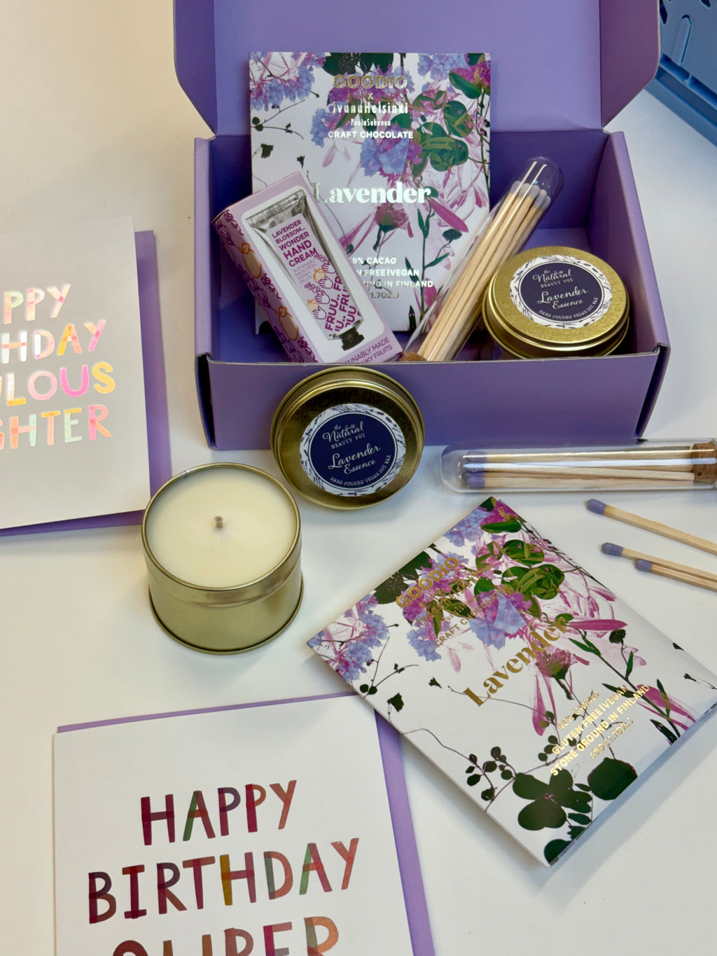 A curated relaxing gift set featuring lavender-themed items including a gold candle, a hand cream, chocolate with match sticks, and a small lavender tin candle, arranged on a white surface.