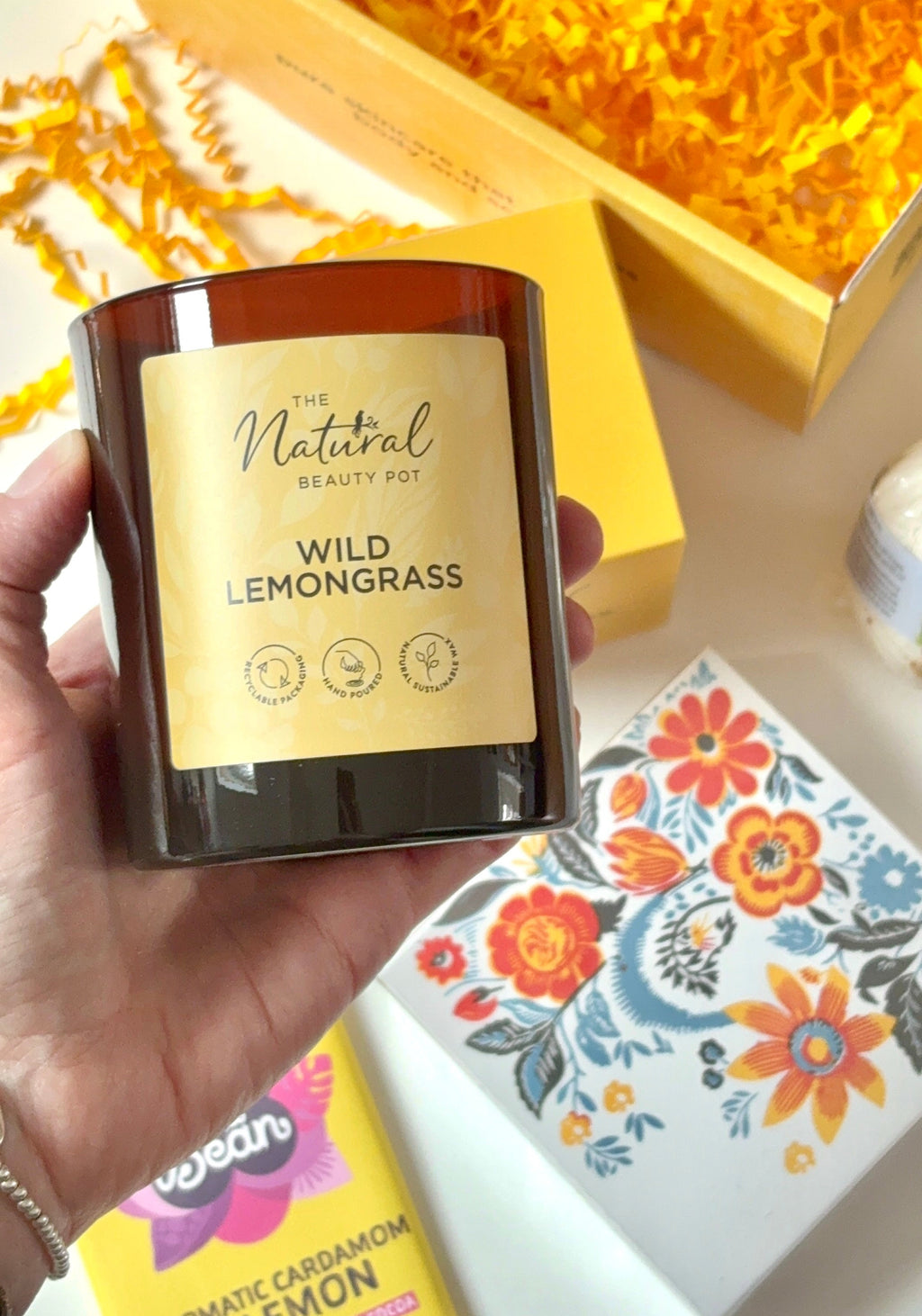 Hand holding a candle labeled 'Wild Lemongrass' with a yellow gift box and floral matches