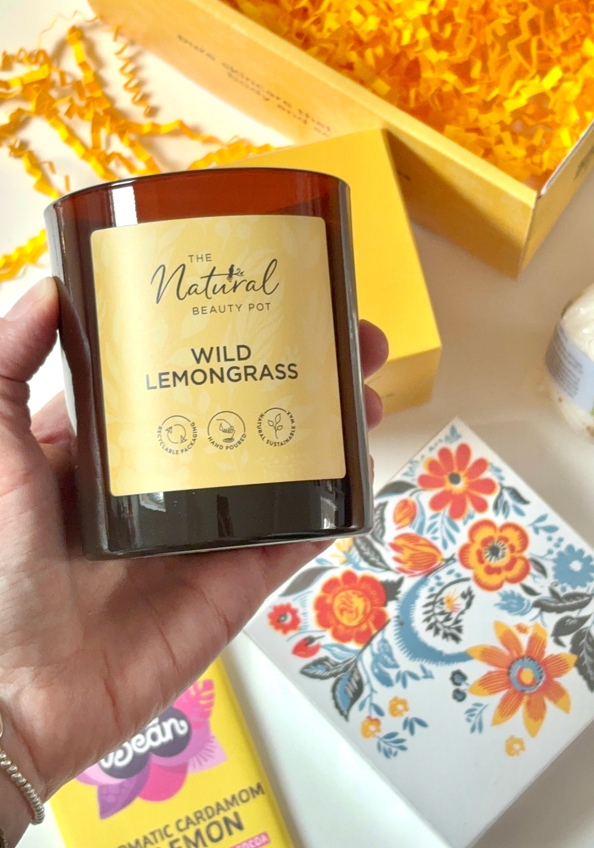 Hand holding a candle labeled 'Wild Lemongrass' with a yellow gift box and floral matches