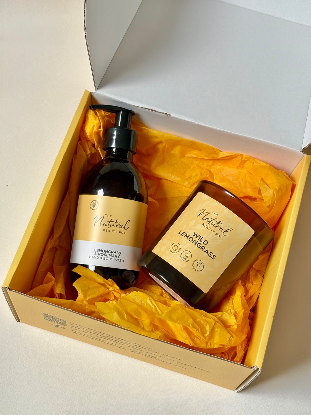 A gift set from Natural Beauty featuring Lemongrass & Rosemary hand wash and a matching Wild Lemongrass candle in a glass jar, both with yellow labels, nestled in bright yellow tissue paper inside an branded gift box.