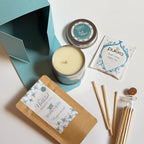 A gift set arranged on a white surface, including a soy candle tin, peppermint bath salts, 'Pukka tea', snatches and a glass tube with more wicks, alongside an open bright blue box.