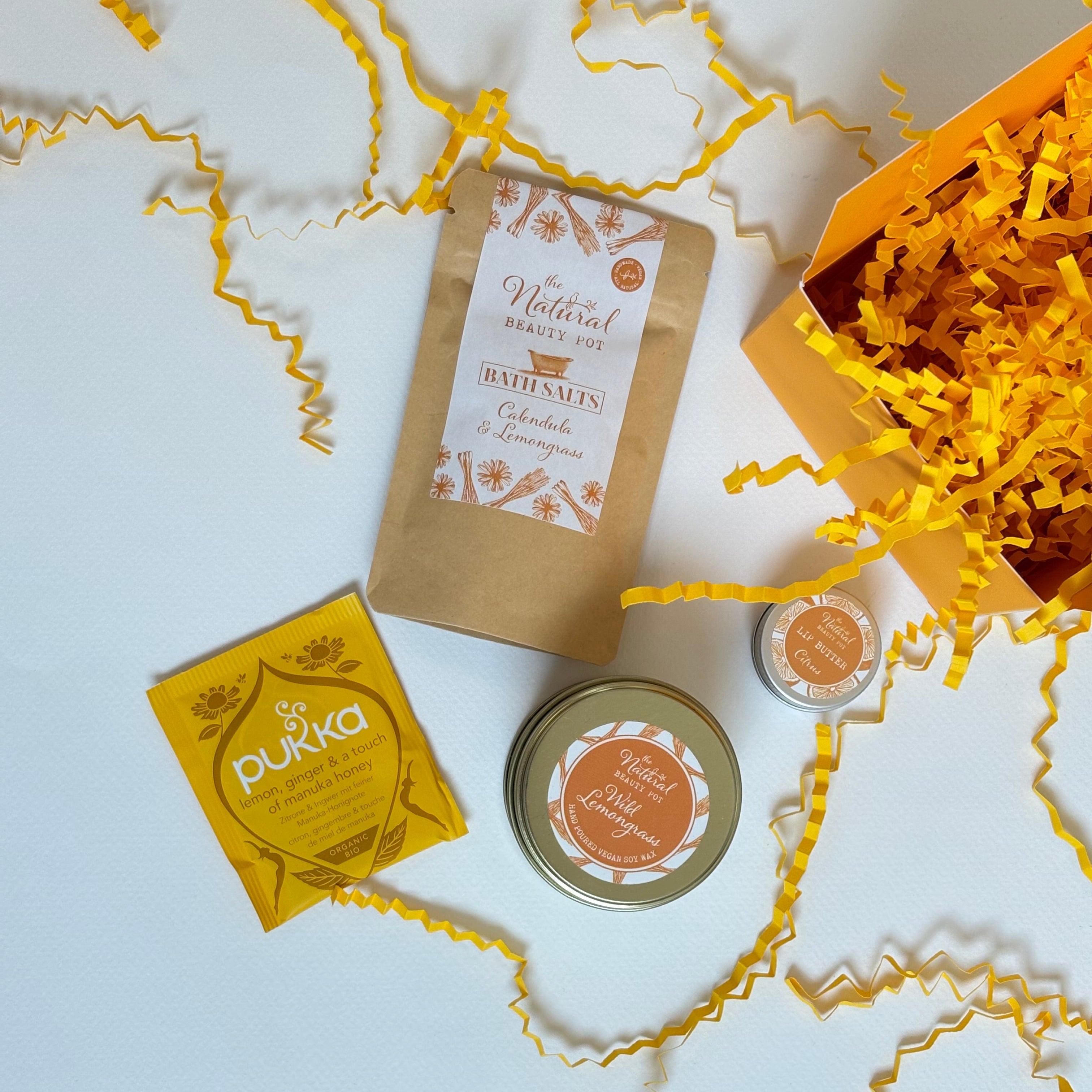 A curated gift set featuring a yellow gift box, Lemongrass & calendula Bath Salts sachet, a yellow Pukha tea, a small round lemongrass candle tin with yellow shredded paper.