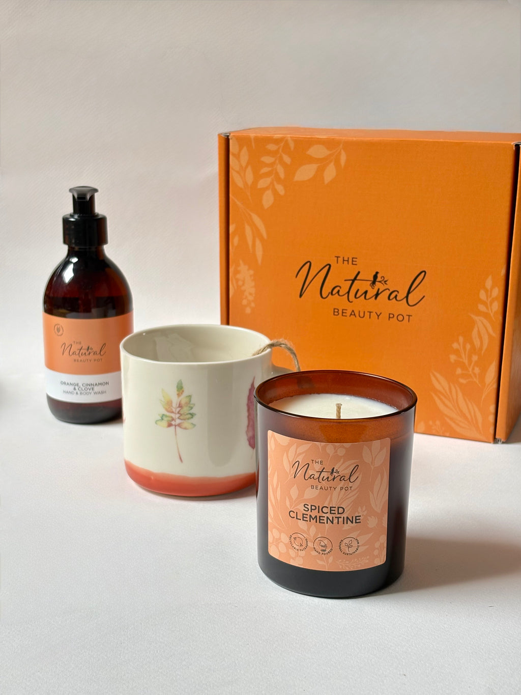 A curated gift set featuring a ceramic mug with a leaf design, a scented candle in an amber glass jar, and a matching amber pump bottle, all arranged in front of an orange branded box on a light neutral background.