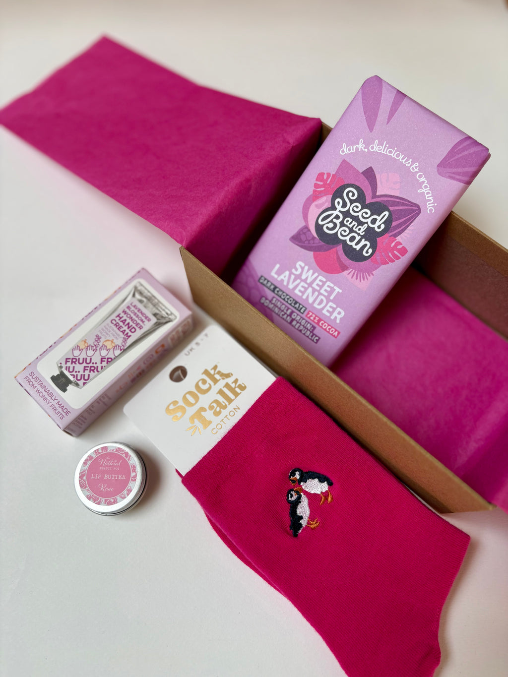 Pink socks with a puffin design, a small lip butter, lavender hand cream and a Seed Bean chocolate  on a white background.