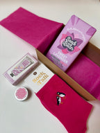Pink socks with a puffin design, a small lip butter, lavender hand cream and a Seed Bean chocolate  on a white background.