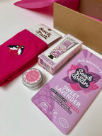 Pink socks with penguin design, lavender hand cream small lip balm tin, and Sweet Lavender chocolate bar on a white surface.