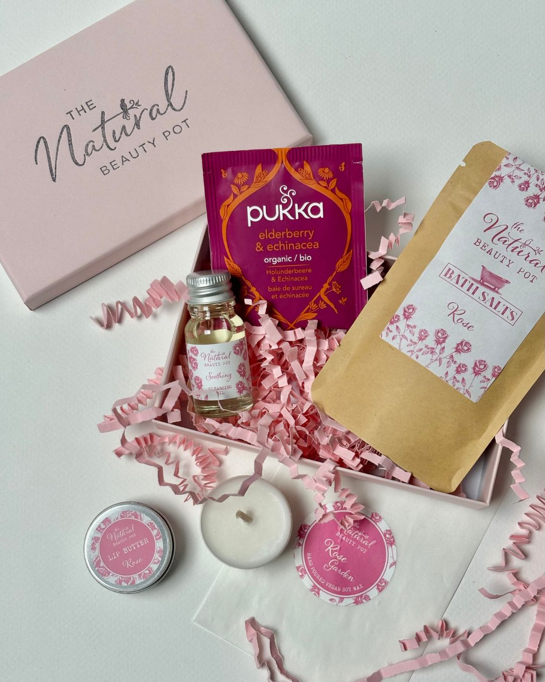 Collection of relaxing rose natural beauty products including tea light candles, tea, bath salts, lip balm and cleansing oil in a matching pink box