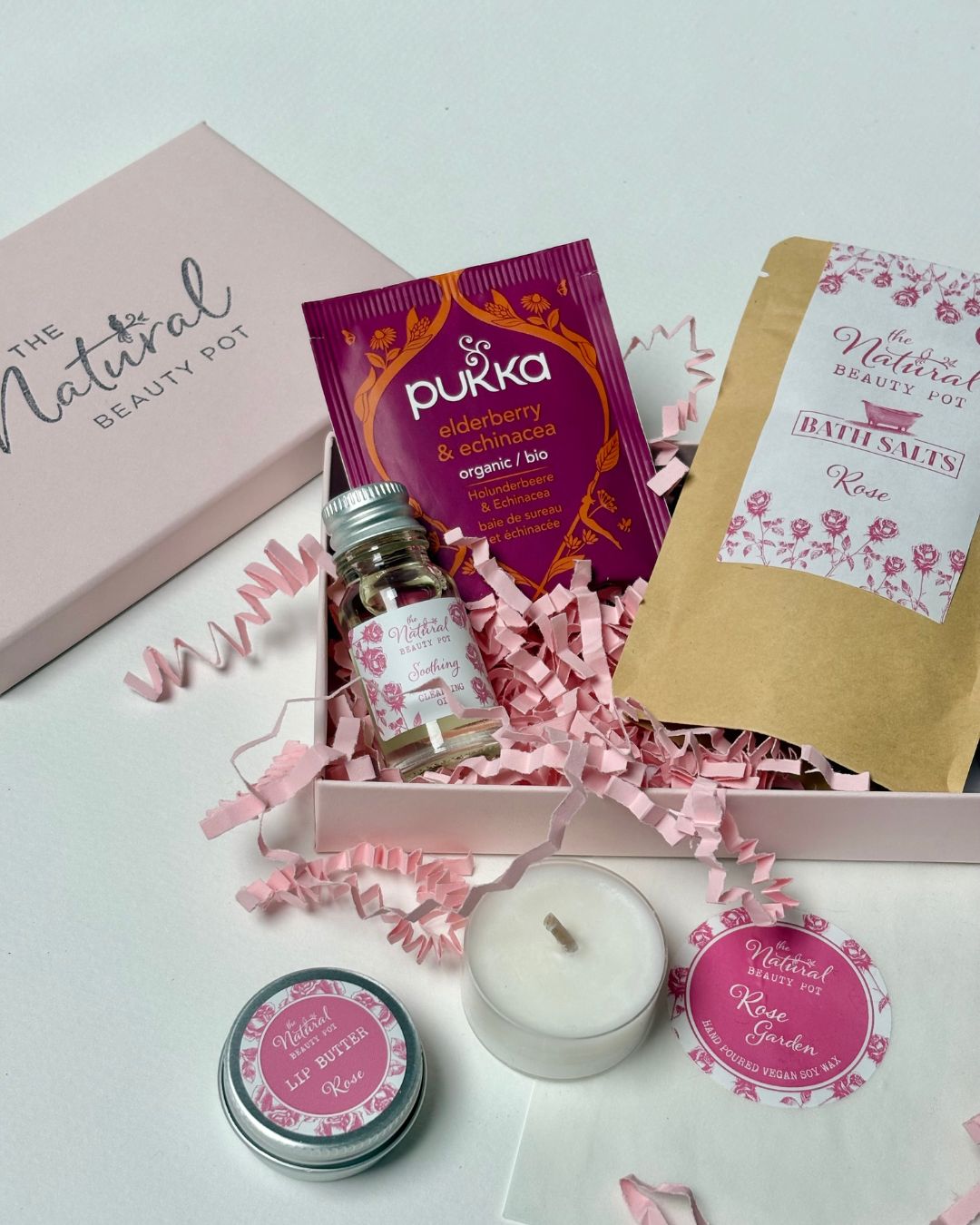 Collection of natural rose well being products including tea light candles, tea, bath salts, lip balm and skincare items on a light background with decorative ribbons.