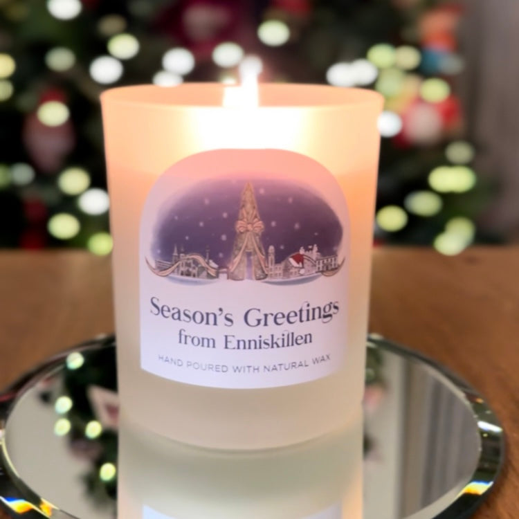 Candle with 'Season's Greetings from Enniskillen' label on a reflective surface with blurred festive background