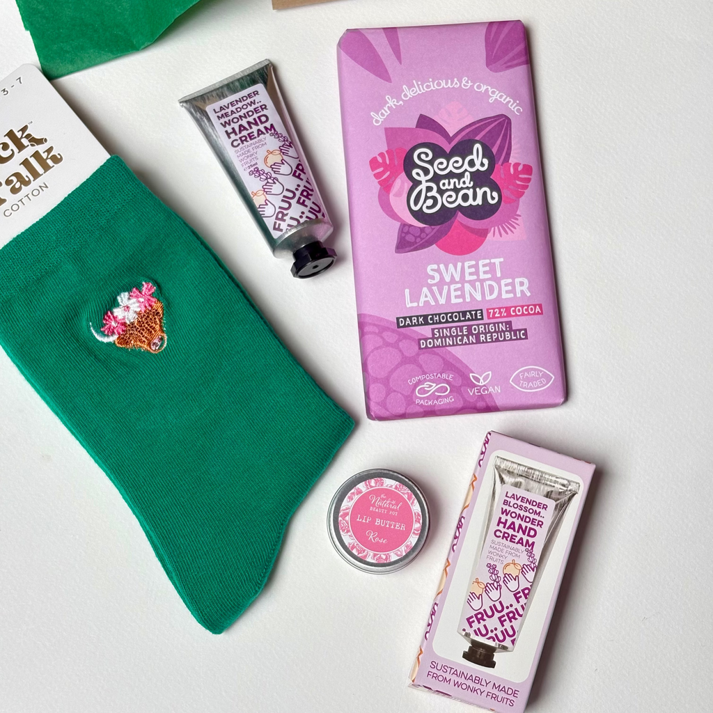 A curated gift set arranged on a white surface, including  green bamboo socks, a purple bar of Seed and Bean Sweet Lavender chocolate, a lavender hand cream tube, rose lip butter, and a pink box, alongside green tissue paper and a cardboard gift box.