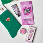 A curated gift set arranged on a white surface, including  green bamboo socks, a purple bar of Seed and Bean Sweet Lavender chocolate, a lavender hand cream tube, rose lip butter, and a pink box, alongside green tissue paper and a cardboard gift box.