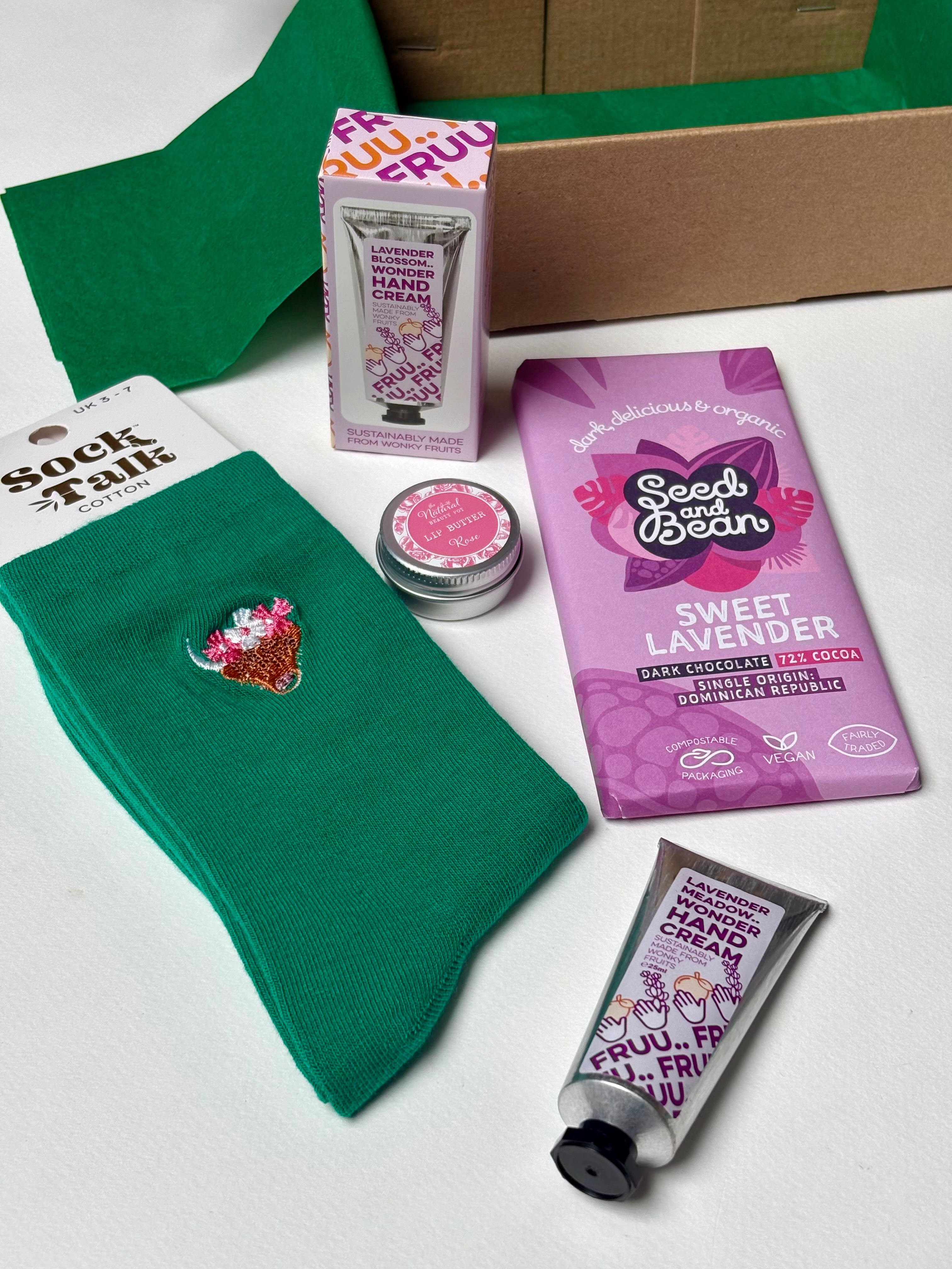 A curated gift set featuring a pair of green highland cow socks, a bar of Seed on Bean Sweet Lavender chocolate, a tube of Fruu hand cream, a rose lip balm, and a box of Fruit of the Loom hand cream