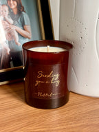 A dark amber glass candle with white wax sits on a wooden surface, featuring a gold foil label that reads 'Sending you a hug' and 'The Natural Beauty Pot'. In the background, a white textured ceramic vase and a framed photo.