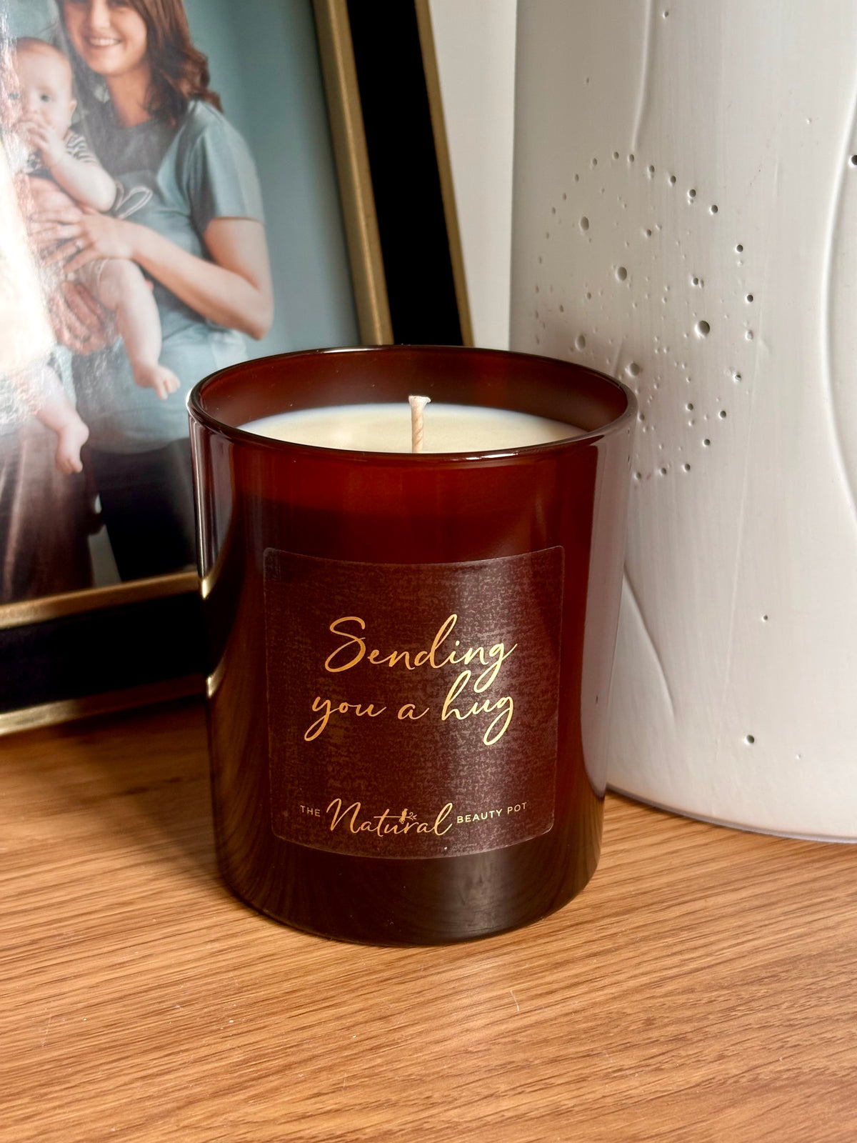 A dark amber glass candle with white wax sits on a wooden surface, featuring a gold foil label that reads 'Sending you a hug' and 'The Natural Beauty Pot'. In the background, a white textured ceramic vase and a framed photo.