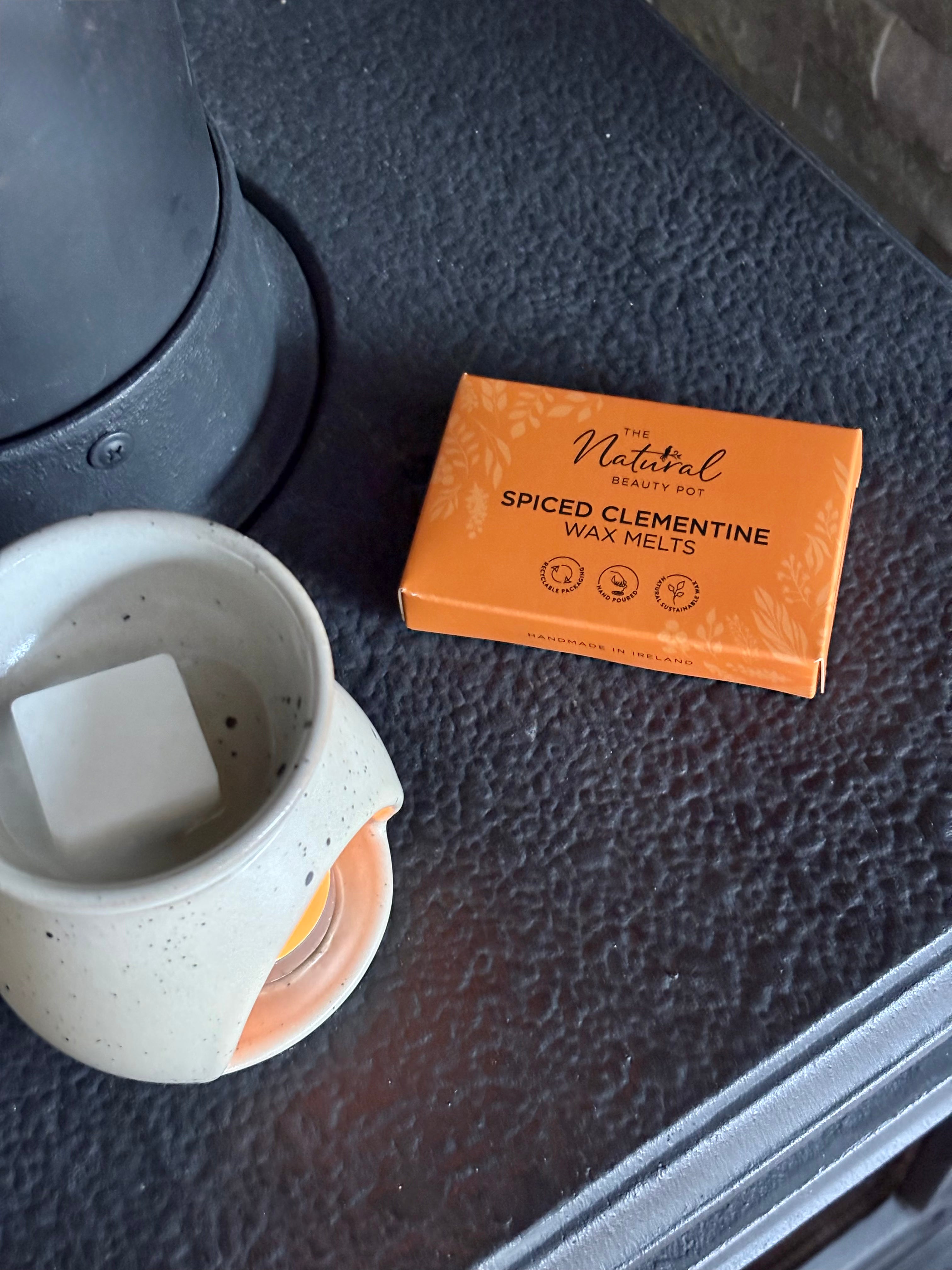 A packet The Natural Beauty Pot Spiced Clementine wax melts sits on a dark textured surface next to a ceramic warmer with a wax melt cube inside.