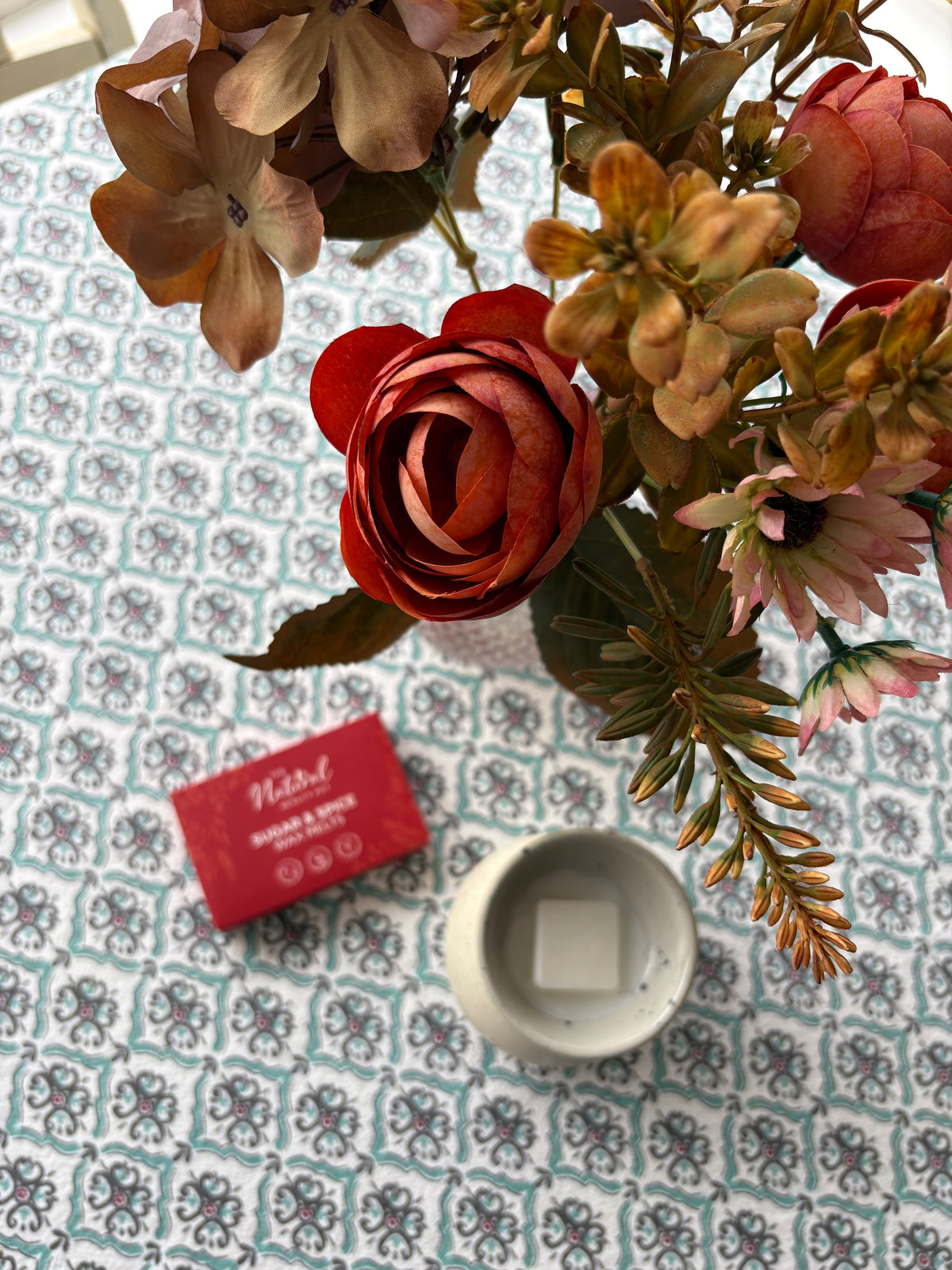 An artificial floral arrangement in a vase on a patterned tablecloth, with a red box labeled The Natural Beauty Pot Sugar & Spie Wax Melts' and a white ceramic burner