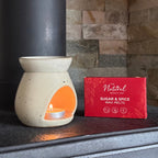 A ceramic wax warmer pot with a lit tea light candle inside, placed on a dark metal surface beside a red box labeled 'The Natural Beauty Pot Sugar & Spice Wax Melts'. The background features a stone fireplace wall.