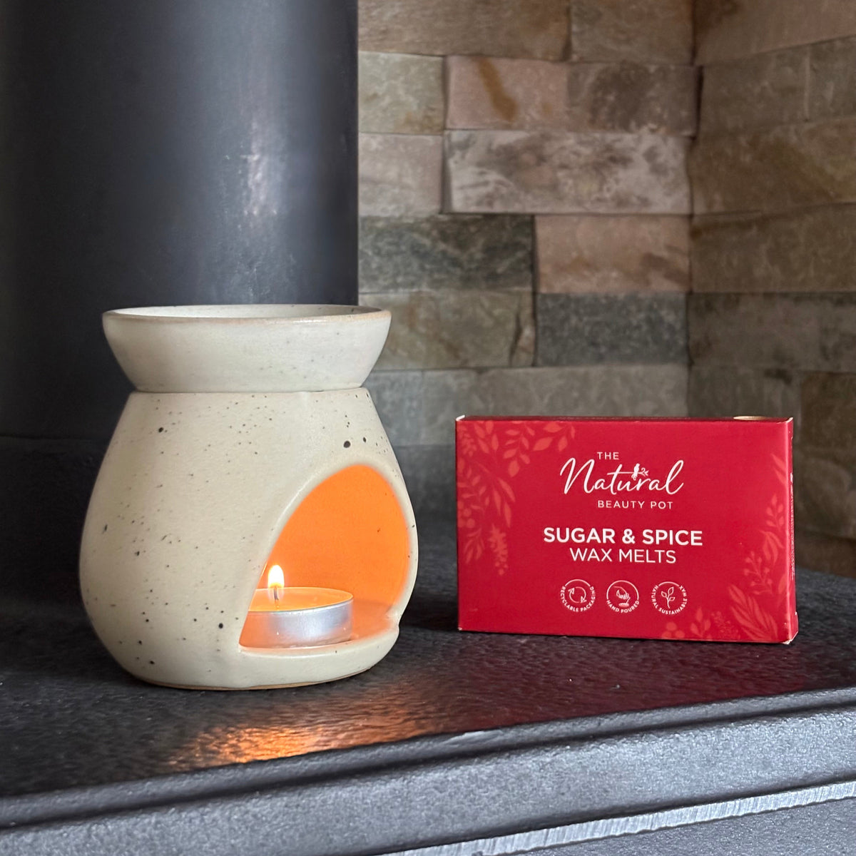 A ceramic wax warmer pot with a lit tea light candle inside, placed on a dark metal surface beside a red box labeled 'The Natural Beauty Pot Sugar & Spice Wax Melts'. The background features a stone fireplace wall.