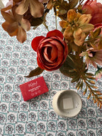 A styled flat-lay of a red wax melts box labeled 'The Natural Beauty Pot Sugar & Spice' next to a small white ceramic wax melt burner containing a white wax melt, arranged with dried flowers on a patterned blue and white tablecloth. 