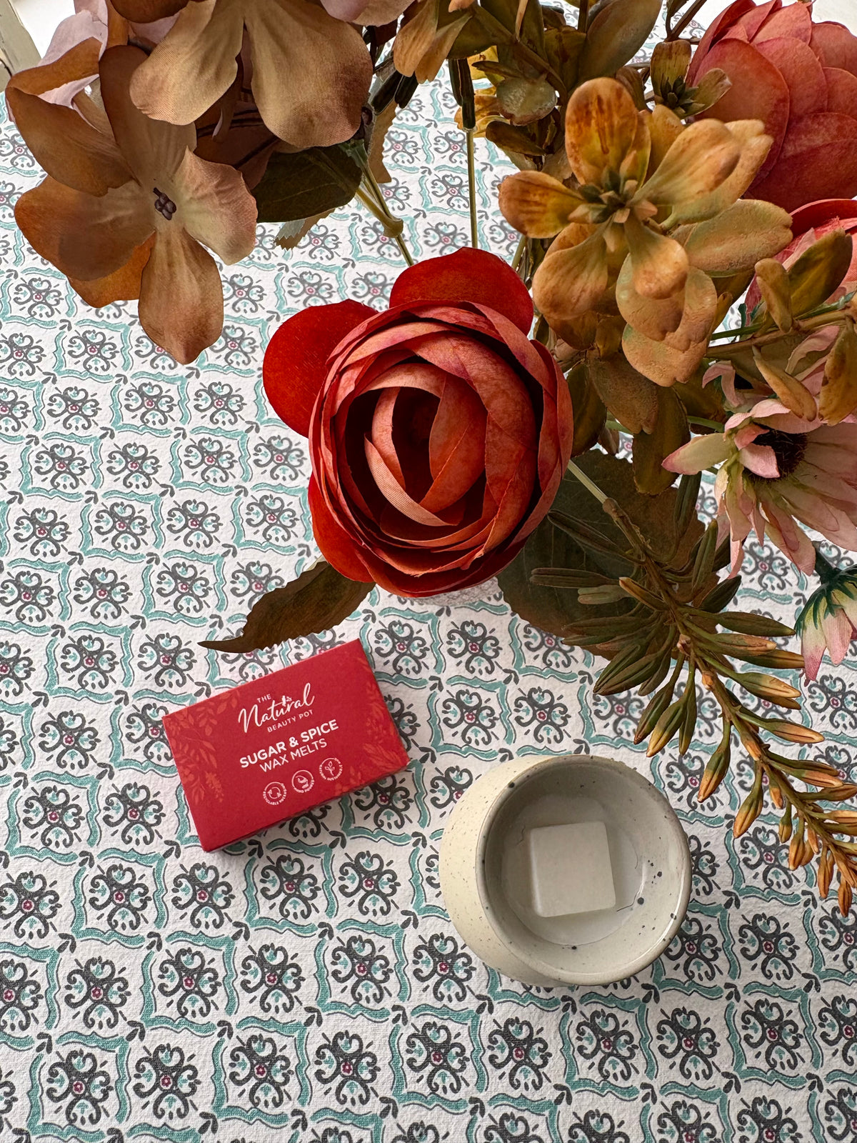 A styled flat-lay of a red wax melts box labeled 'The Natural Beauty Pot Sugar & Spice' next to a small white ceramic wax melt burner containing a white wax melt, arranged with dried flowers on a patterned blue and white tablecloth. 