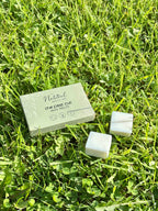 Natural square wax melts  are placed on green grass next to their light sage green packaging box, which reads 'The First Cut Wax Melts' by 'The Natural Beauty Pot".