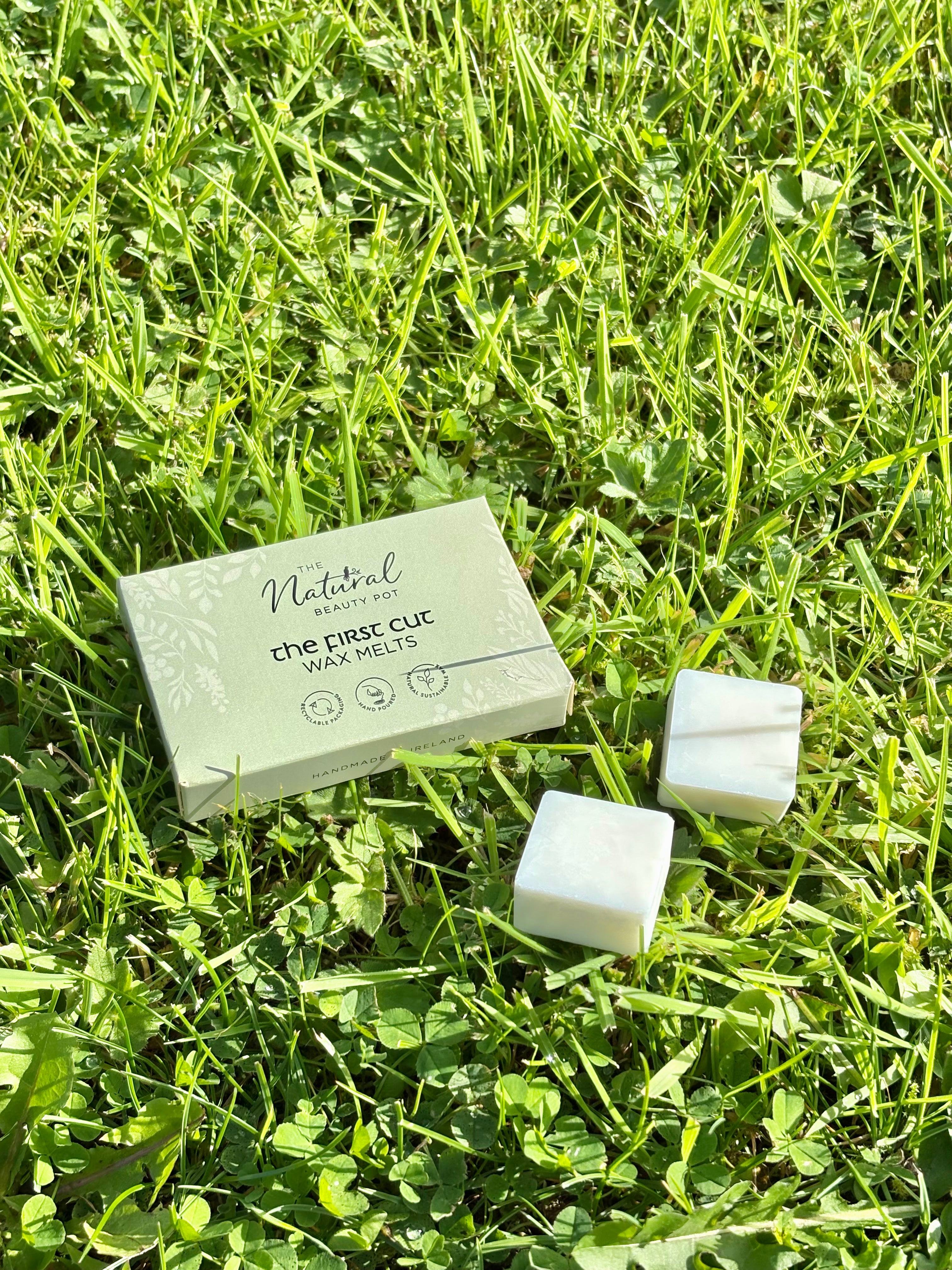 Natural square wax melts  are placed on green grass next to their light sage green packaging box, which reads 'The First Cut Wax Melts' by 'The Natural Beauty Pot".