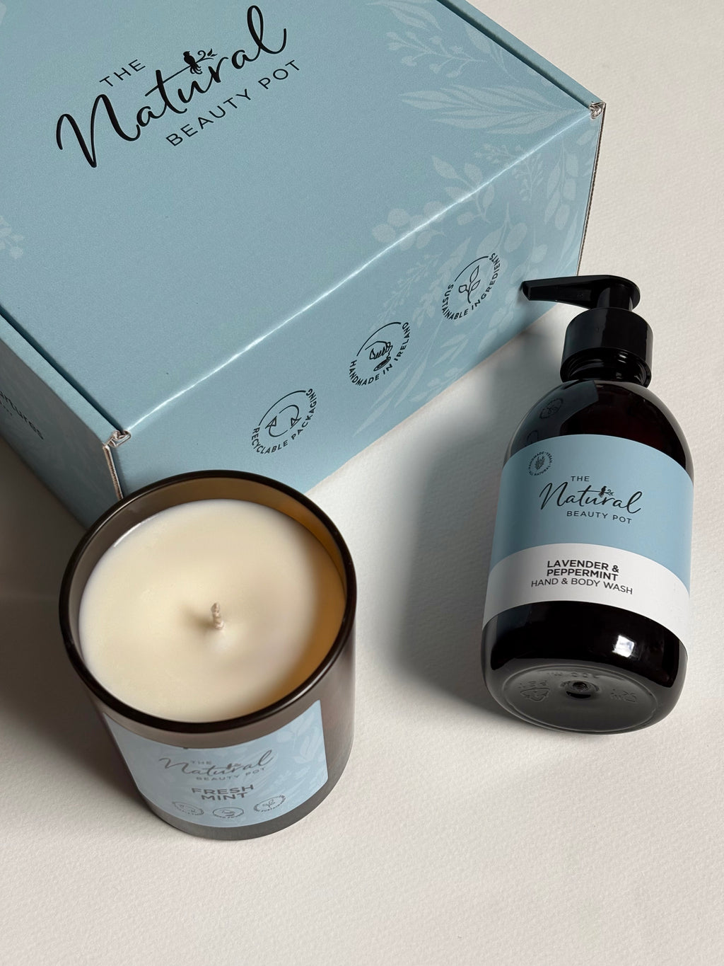 The Natural Beauty Pot hand wash and candle set, featuring a lavender and peppermint hand wash in a dark amber bottle with a black pump, and fresh mint cented candle in a brown glass jar, arranged in front of their light blue branded gift box.