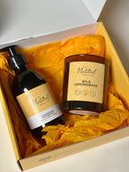 A gift set from Natural Beauty featuring Lemongrass & Rosemary hand wash and a matching Wild Lemongrass candle in a glass jar, both with yellow labels, nestled in bright yellow tissue paper inside an branded gift box.