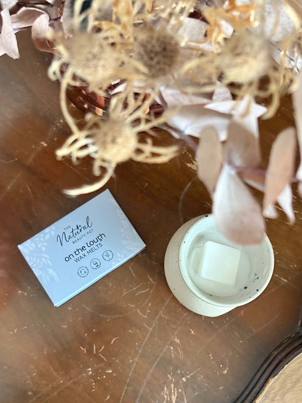 A light blue box labeled 'The Natural  Beauty Pot On the Lough Wax Melts' sits on a wooden table beside a white ceramic wax melt burner holding a square wax melt, with dried flowers in the background.