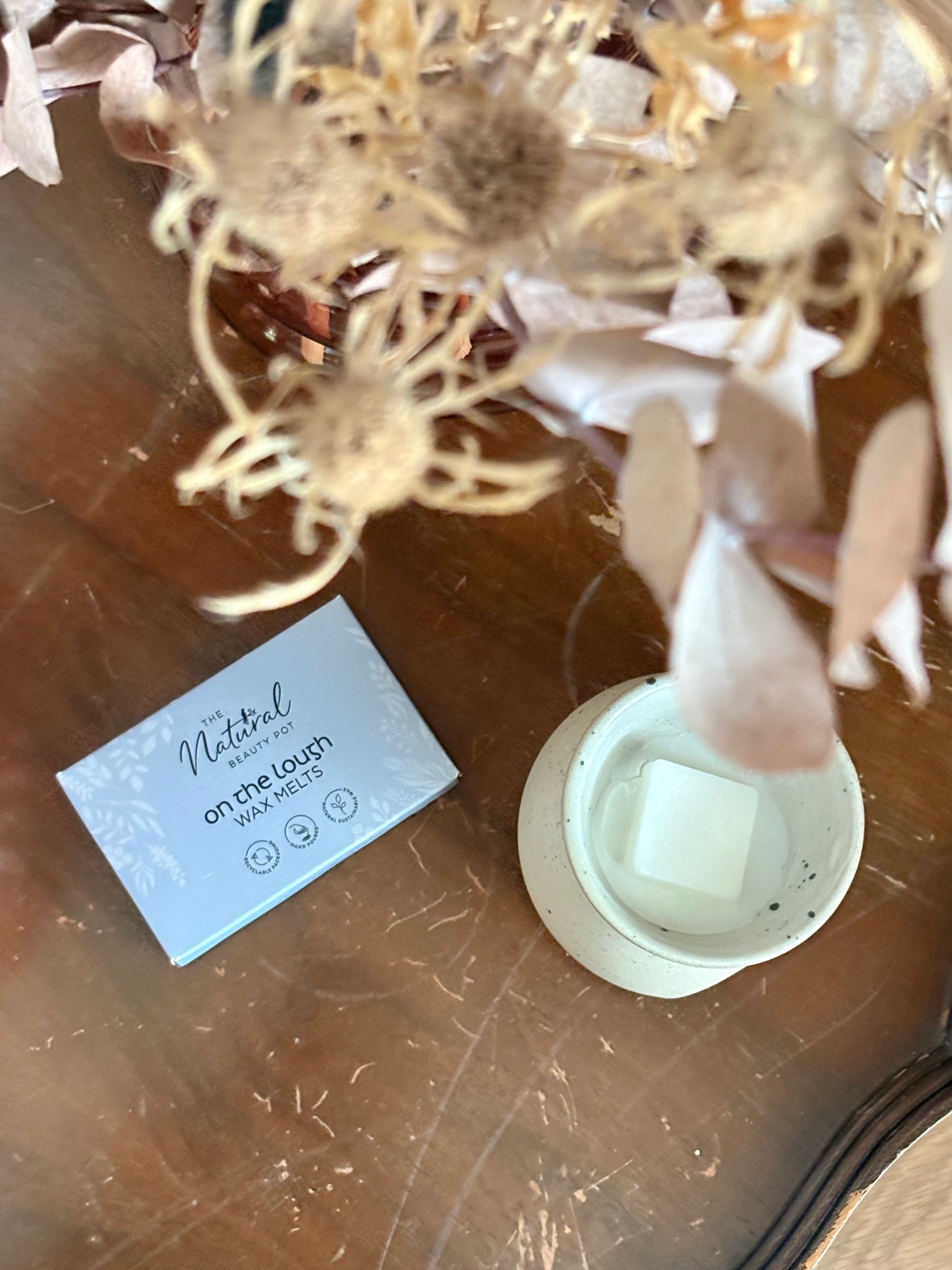 A light blue box labeled 'The Natural  Beauty Pot On the Lough Wax Melts' sits on a wooden table beside a white ceramic wax melt burner holding a square wax melt, with dried flowers in the background.