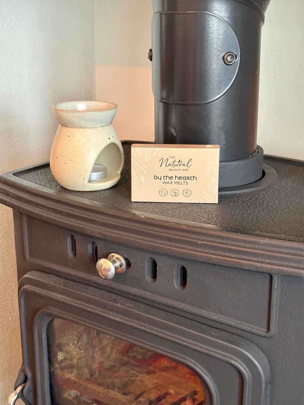 A ceramic scented candle in a small cream-colored holder sits on top of a dark cast iron wood stove next to a metal chimney pipe and a branded card that reads 'Natural by the beach'.