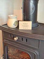 A ceramic scented candle in a small cream-colored holder sits on top of a dark cast iron wood stove next to a metal chimney pipe and a branded card that reads 'Natural by the beach'.